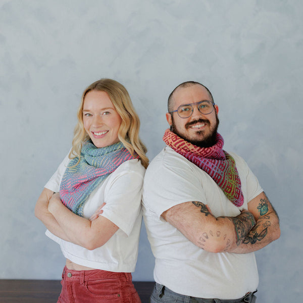 A smiling blonde woman and a bearded man with glasses stand back-to-back against a textured light blue-gray wall, each wearing a colorful hand-knit cowl scarf. The woman's scarf is pastel blue, pink, and purple, while the man's is pink, red, and green.